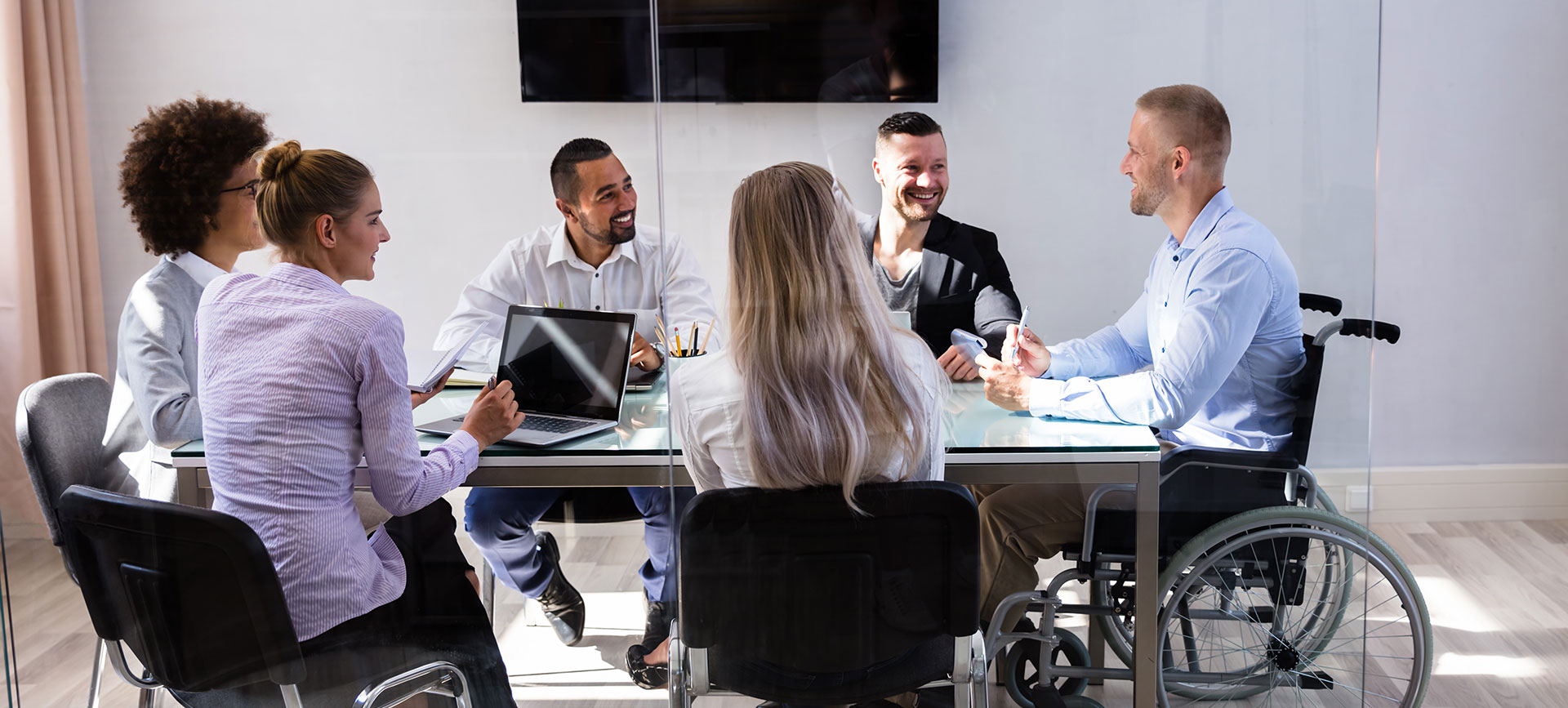6 people sat around meeting table, having discussions. One person is in a wheelchair.